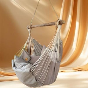 May include: A gray hanging chair with a wooden bar and white rope supports. The chair is filled with gray pillows and a blanket. The background features a soft, warm-toned curtain.