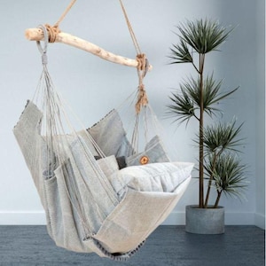 Puede incluir: Una silla hamaca colgante hecha de tela tejida gris claro, suspendida de una rama de madera. La silla está llena de varias almohadas a juego. Una planta decorativa en una maceta gris está en el fondo.