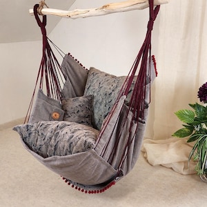 May include: A hanging chair made of gray fabric with deep red rope suspension and matching pom-pom trim. The seat includes two gray faux fur pillows and a small square pillow, all suspended from a light-colored wooden branch with thick rope.