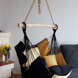 Könnte beinhalten: Ein Hängesessel mit einer Sitzfläche aus schwarzem und senfgelbem Stoff, der an schwarzen Seilen von einem hellen Holzstab herabhängt. Der Sessel enthält zwei passende senfgelbe quadratische Kissen und ein schwarzes Sitzkissen, die an einem dicken Seil hängen.