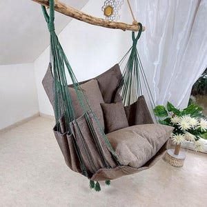 May include: A hanging chair with a brown fabric seat and back, supported by green rope and a natural wooden beam. The chair is filled with several brown pillows and cushions, creating a cozy seating area. The design is rustic and inviting.
