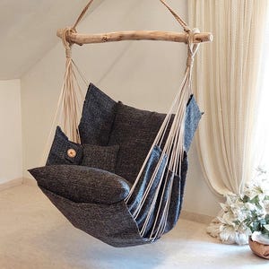 May include: A hanging chair featuring a dark charcoal gray fabric seat with integrated cushions, suspended by thick cream-colored ropes from a natural light-colored wooden branch. The chair includes a small square side pocket with a circular logo patch.