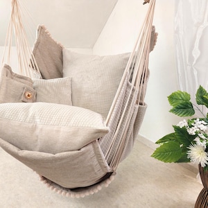 Puede incluir: Una silla colgante de color beige con tela de textura tipo gofre, varios cojines mullidos y cuerdas de suspensión de color crema. La silla está adornada con un borde de pequeños pompones en la parte inferior y una etiqueta de madera.