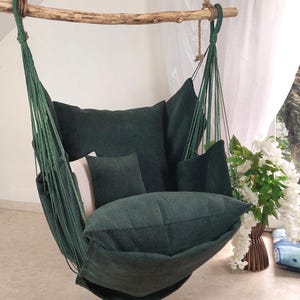 May include: A dark green hanging chair suspended from a natural wooden branch by thick green ropes. The chair features a large back cushion, a smaller square cushion, and a wide seat cushion, all in a matching dark green textured fabric.