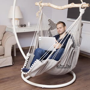 May include: A beige hanging hammock chair with a white metal stand and a wooden spreader bar. The fabric features a textured pattern and a row of white pom-poms along the edge. The text "aix.lv" is printed in black on the lower side of the fabric.