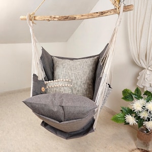 May include: A hanging chair made of dark grey fabric suspended from a natural wooden branch by thick white ropes. The chair includes a dark grey seat cushion and a light grey textured back cushion decorated with a row of small white pom-poms.