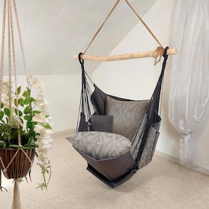 Puede incluir: Una silla colgante con asiento de tela gris oscuro suspendido por cuerdas negras de una barra de madera clara. La silla incluye un cojín de respaldo grande y un cojín de asiento, ambos decorados con un sutil patrón ikat en gris y blanco.
