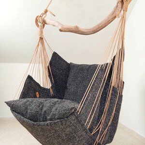 May include: A hanging chair featuring a dark charcoal grey textured fabric seat and backrest, suspended by light beige ropes from a curved natural wooden branch. The chair includes a plush seat cushion and a matching back cushion for support.