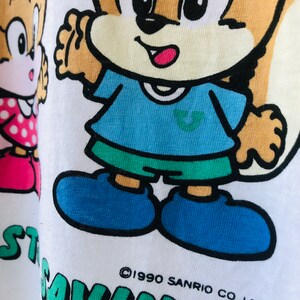 Puede incluir: Camiseta blanca con una ardilla de dibujos animados con una camisa azul y pantalones cortos verdes. La ardilla tiene una cara bronceada y ojos negros. El texto "START SAVINGS" est&aacute; impreso en verde. El copyright dice "&copy;1990 SANRIO CO., LTD."