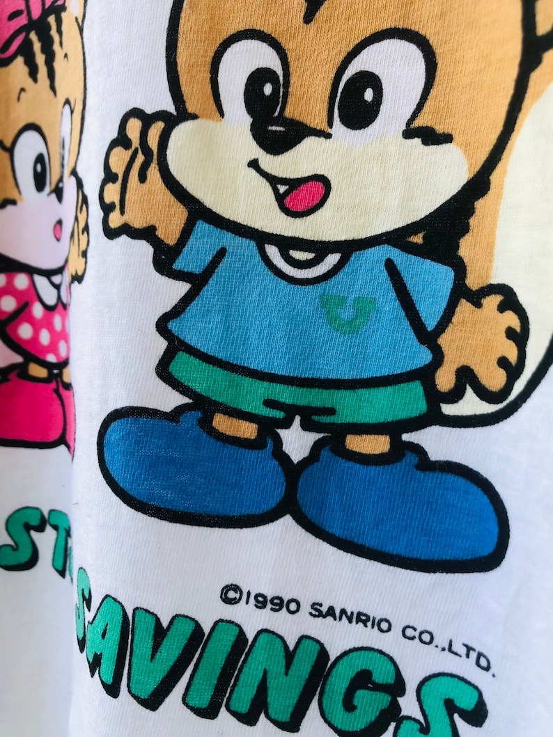 Puede incluir: Camiseta blanca con una ardilla de dibujos animados con una camisa azul y pantalones cortos verdes. La ardilla tiene una cara bronceada y ojos negros. El texto "START SAVINGS" est&aacute; impreso en verde. El copyright dice "&copy;1990 SANRIO CO., LTD."