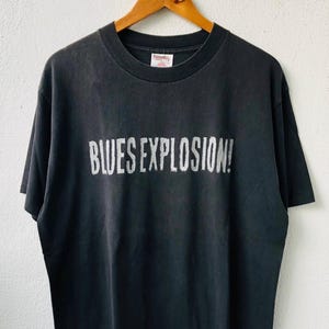 May include: Black t-shirt with the text "BLUES EXPLOSION!" in distressed white lettering. The shirt is hanging on a wooden hanger. The t-shirt has a crew neck and short sleeves.