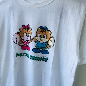 Puede incluir: Camiseta blanca con un dibujo animado de dos personajes parecidos a ardillas. Uno lleva un lazo y un vestido rosa, el otro un gorro y una camisa azules. Las palabras "POSTAL SAVINGS" est&aacute;n impresas en verde debajo.