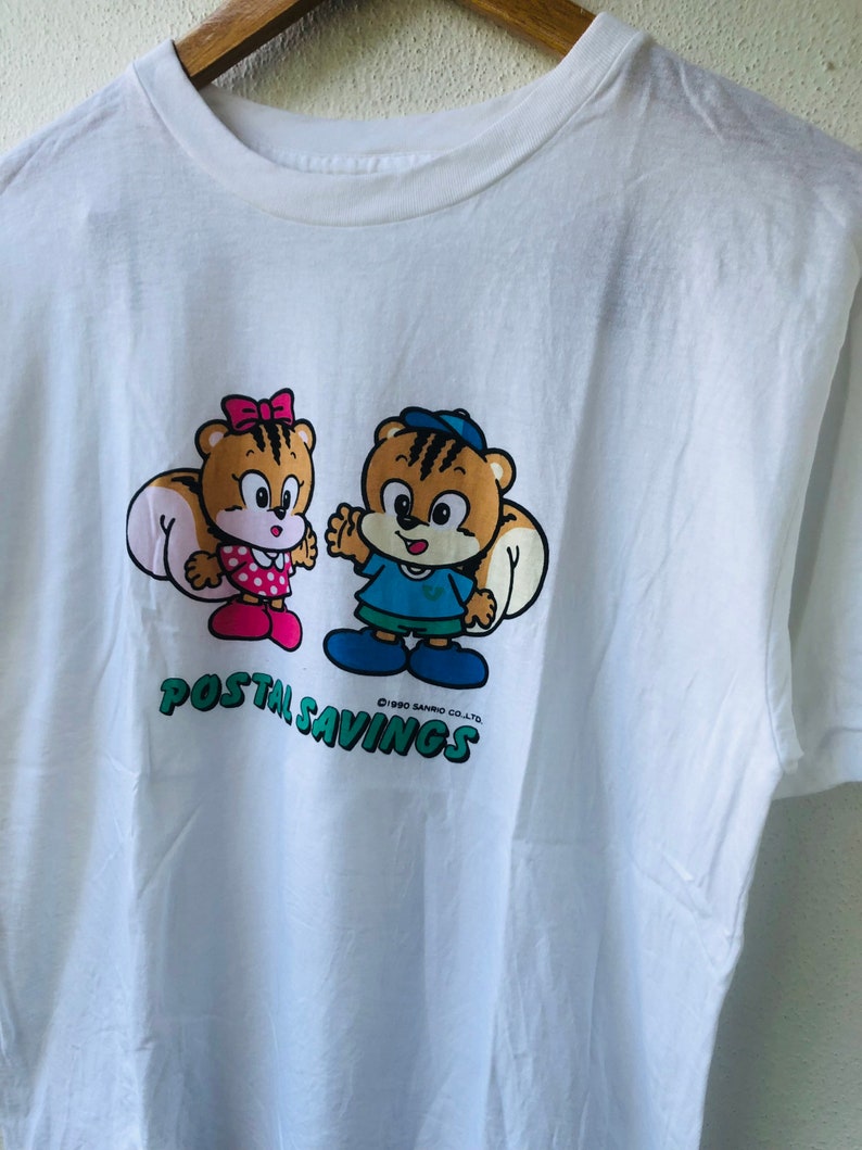 Puede incluir: Camiseta blanca con un dibujo animado de dos personajes parecidos a ardillas. Uno lleva un lazo y un vestido rosa, el otro un gorro y una camisa azules. Las palabras "POSTAL SAVINGS" est&aacute;n impresas en verde debajo.