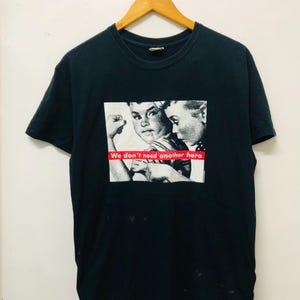 May include: Black t-shirt with a graphic print of two children. The graphic features a black and white image of a boy with his fist raised and a girl with her arm around him. The text "We don't need another hero" is printed in red across the bottom of the graphic.
