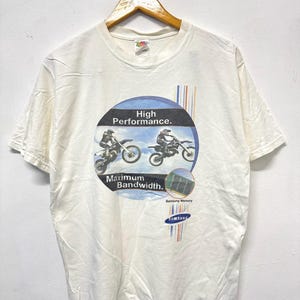 May include: A white short-sleeve t-shirt featuring a graphic of two motocross riders in mid-air. The text reads High Performance. Maximum Bandwidth. Samsung Memory and Samsung. The design includes a blue circle, vertical stripes, and a small image of a memory module.