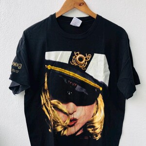 以下が含まれることがあります： 黒い帽子とサングラスをかけた女性のグラフィックが特徴の黒いTシャツ。女性はブロンドの髪と赤いリップスティックをしています。シャツは木製のハンガーに吊るされています。