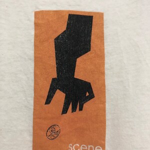 May include: A white t-shirt with a brown rectangle printed on the front. The rectangle contains a black abstract design and the word "scene" printed in white.