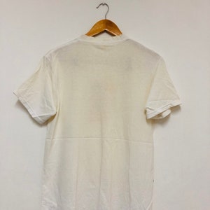 May include: A white t-shirt hanging on a wooden hanger against a white wall.