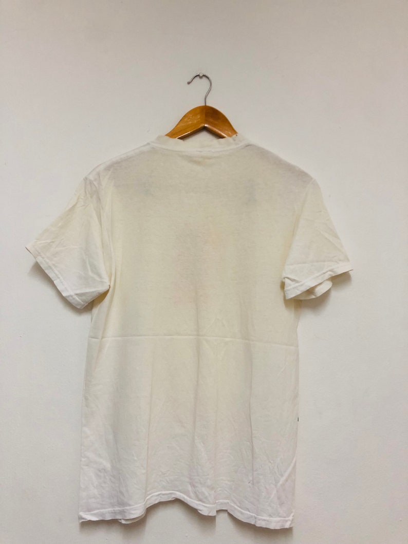 May include: A white t-shirt hanging on a wooden hanger against a white wall.