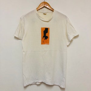 May include: White t-shirt with a black and orange graphic of a hand holding a chair. The graphic is in a square with the word "scene" below it.