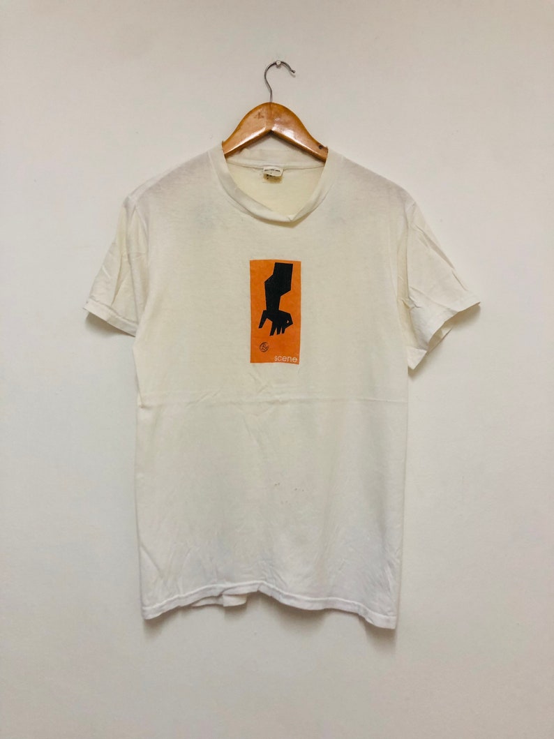 May include: White t-shirt with a black and orange graphic of a hand holding a chair. The graphic is in a square with the word "scene" below it.