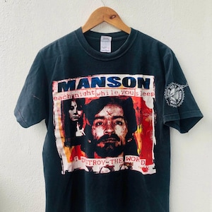 May include: Black t-shirt featuring a graphic design with the word "MANSON" in blue and white. The design includes a portrait and the text "each night while you sleep I DESTROY THE WORLD". A circular graphic is on the left sleeve.
