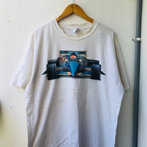 Puede incluir: Camiseta blanca con un gráfico de un coche de carreras azul en una pista. El coche tiene un conductor en la cabina y el texto "Miller" es visible en el coche.