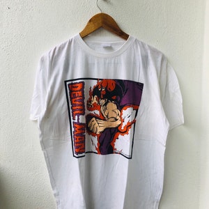 Puede incluir: Camiseta blanca con un estampado gráfico de un personaje de dibujos animados con cuernos rojos y una capa morada. El texto "Devil Man" está impreso verticalmente en el lado izquierdo del gráfico.