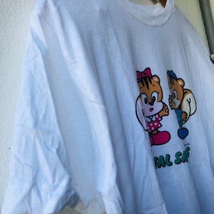 Puede incluir: Camiseta blanca con una ilustraci&oacute;n de dibujos animados de dos personajes con forma de oso. Uno lleva un lazo y un vestido rosa, el otro un sombrero y un pantal&oacute;n azules. El texto "TOTAL SAFE" est&aacute; impreso debajo.