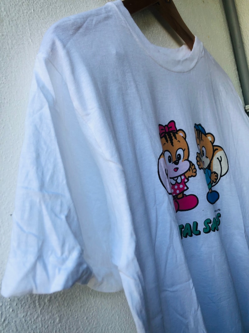 Puede incluir: Camiseta blanca con una ilustraci&oacute;n de dibujos animados de dos personajes con forma de oso. Uno lleva un lazo y un vestido rosa, el otro un sombrero y un pantal&oacute;n azules. El texto "TOTAL SAFE" est&aacute; impreso debajo.