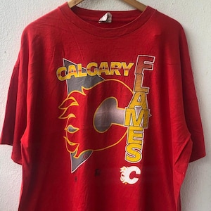 May include: Red t-shirt featuring the Calgary Flames logo. The logo includes the word "CALGARY" above a stylized "C" with flames, and the word "FLAMES" vertically. The shirt is hanging on a wooden hanger.