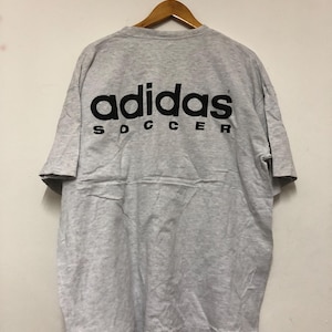 May include: A heather gray Adidas Soccer t-shirt hangs on a wooden hanger. The back of the shirt features the Adidas logo in black block letters above the word "SOCCER". The shirt is made of a soft, comfortable material.