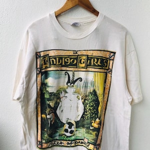 May include: White t-shirt featuring a colorful graphic of the Indigo Girls band. The design includes the band's name, a surreal illustration, and the text "rites of passage". The shirt is hanging on a wooden hanger.