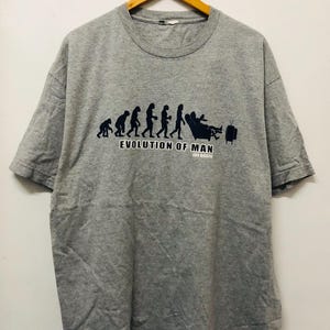 May include: A gray t-shirt with a black and white graphic of the evolution of man, ending with a man sitting in a recliner watching television. The text "EVOLUTION OF MAN" is printed below the graphic.