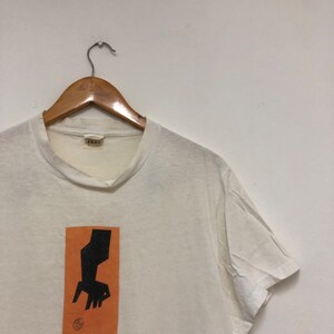 May include: White t-shirt with an orange rectangle graphic featuring a black silhouette of a hand reaching down towards a circle. The text "scene" is printed below the graphic.