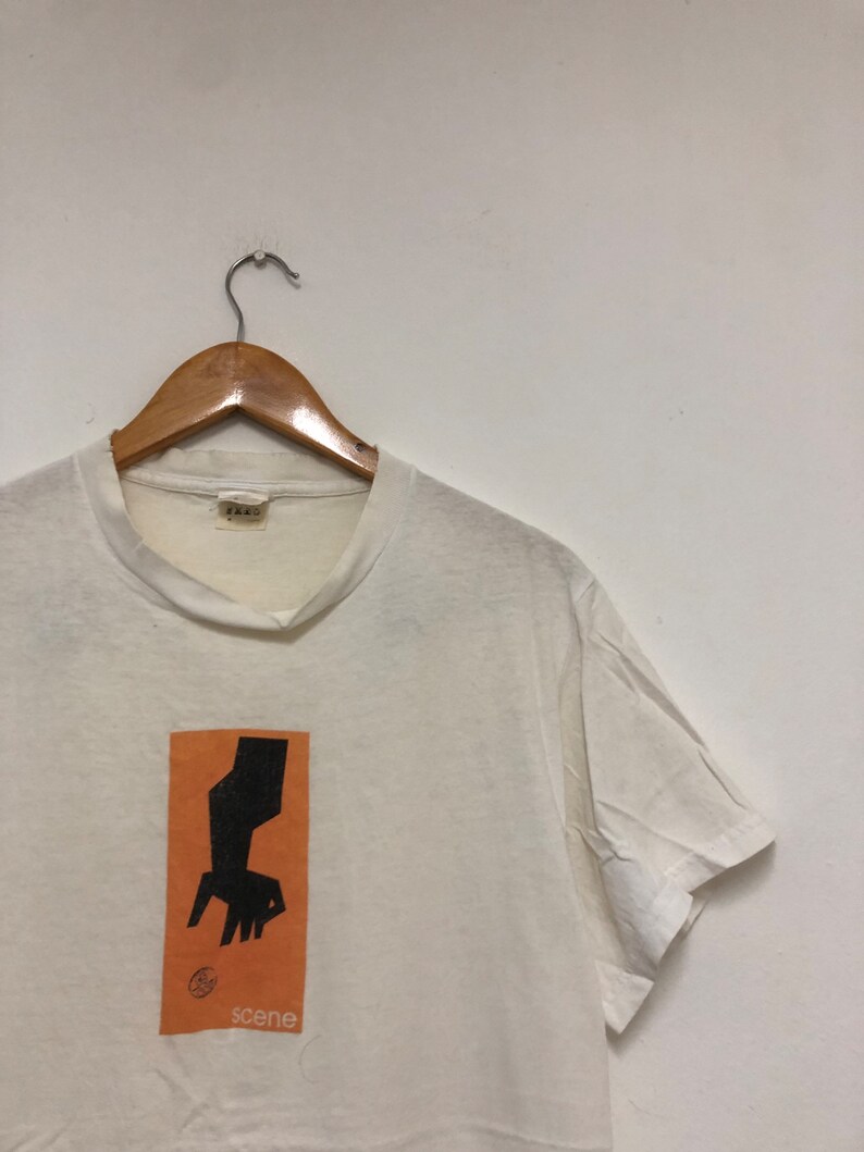 May include: White t-shirt with an orange rectangle graphic featuring a black silhouette of a hand reaching down towards a circle. The text "scene" is printed below the graphic.