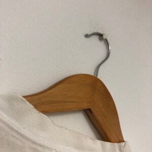 May include: White t-shirt hanging on a wooden hanger. The t-shirt has a crew neck and is made of a soft, lightweight fabric.