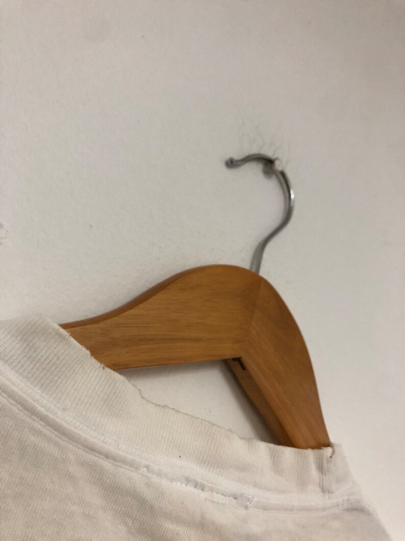 May include: White t-shirt hanging on a wooden hanger. The t-shirt has a crew neck and is made of a soft, lightweight fabric.
