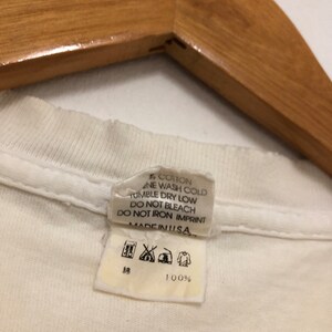 May include: White cotton t-shirt tag with care instructions. The tag says "100% Cotton", "Machine Wash Cold", "Tumble Dry Low", "Do Not Bleach", "Do Not Iron Imprint", and "Made in USA".