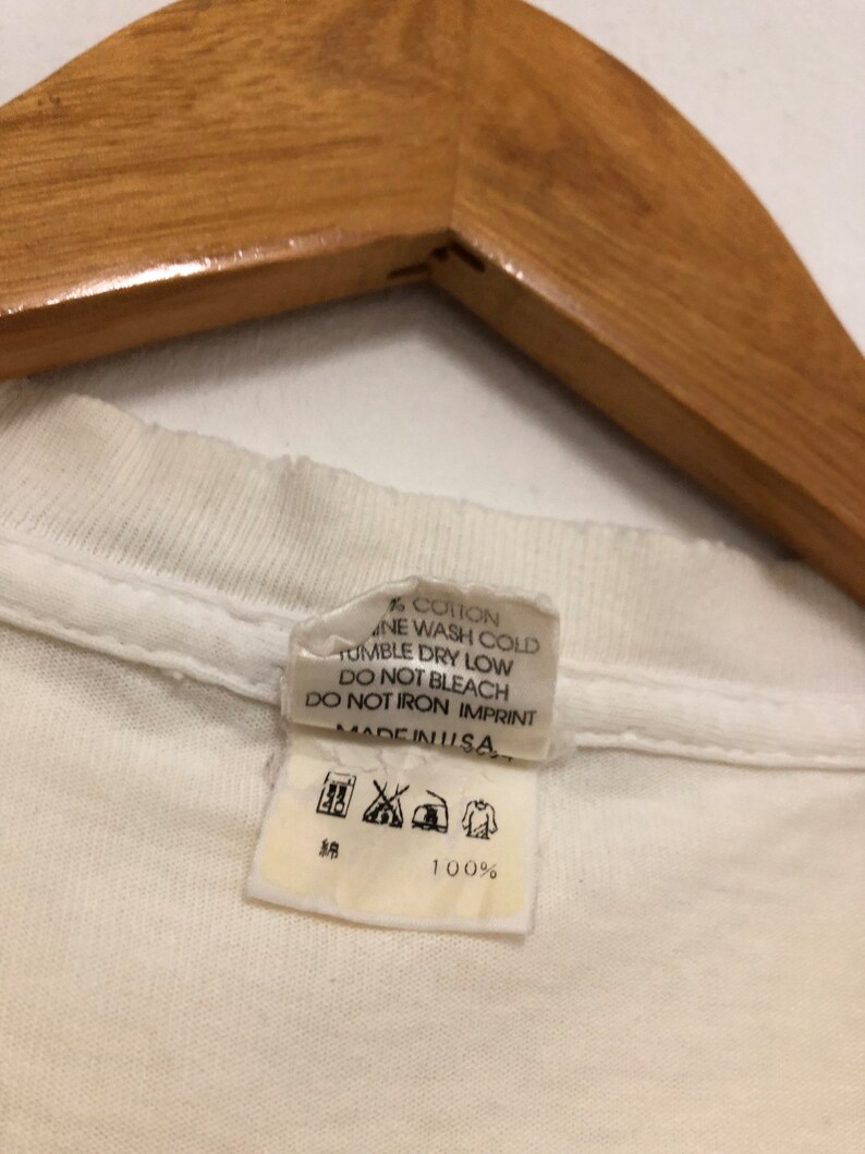 May include: White cotton t-shirt tag with care instructions. The tag says "100% Cotton", "Machine Wash Cold", "Tumble Dry Low", "Do Not Bleach", "Do Not Iron Imprint", and "Made in USA".
