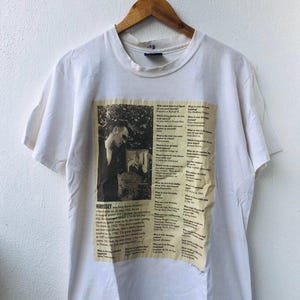 Puede incluir: Camiseta blanca con estampado de periódico que presenta una imagen en blanco y negro de Morrissey. El texto incluye el nombre "Morrissey" y varias preguntas. La camiseta está colgada en una percha de madera contra una pared blanca.