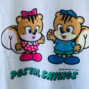 Puede incluir: Camiseta blanca con dos ardillas de dibujos animados. Una ardilla lleva un lazo rosa y un vestido de lunares, la otra una gorra y una camisa azules. El texto verde dice "POSTAL SAVINGS", con "&copy;1990 SANRIO CO.,LTD." debajo.