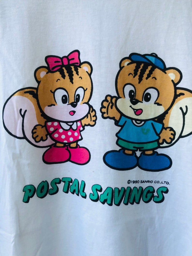Puede incluir: Camiseta blanca con dos ardillas de dibujos animados. Una ardilla lleva un lazo rosa y un vestido de lunares, la otra una gorra y una camisa azules. El texto verde dice "POSTAL SAVINGS", con "&copy;1990 SANRIO CO.,LTD." debajo.