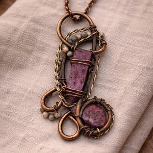 May include: A copper wire-wrapped pendant featuring a central rectangular purple stone and a smaller round purple stone. The design incorporates twisted copper wire accents and small, light-colored spherical beads integrated into the metal frame.