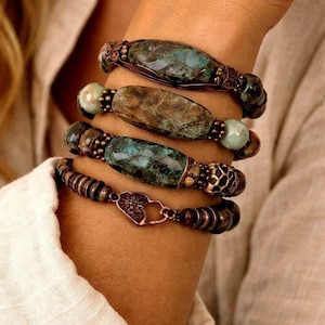 May include: Four stacked bracelets with natural stone beads in shades of green, brown, and copper. The bracelets feature various bead shapes and sizes, with intricate metalwork and a heart-shaped clasp on the bottom bracelet.