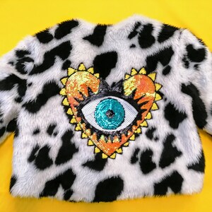 May include: A faux fur jacket with a black and white cow print. The jacket features a sequined heart-shaped patch with an eye design in the centre. The eye is turquoise, and the heart has orange and yellow sequins.