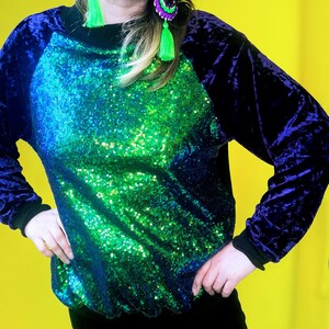 May include: A green and blue sequined top with a purple velvet raglan sleeve. The top has a round neckline.