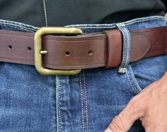 May include: A dark brown leather belt with a rectangular brass buckle, worn over blue denim jeans and a black button-down shirt. A circular brown graphic overlay on the left side contains the white text: Leather balm included with your order.