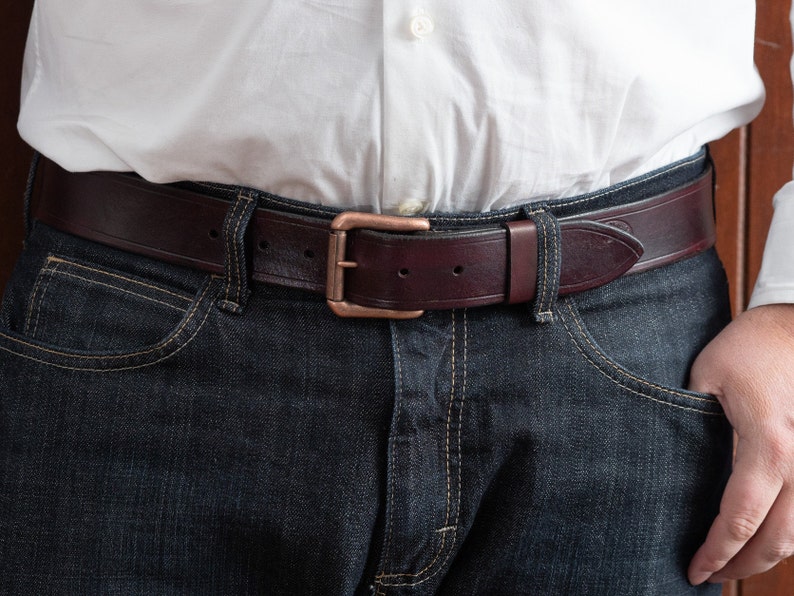 May include: A dark brown leather belt with a copper buckle. The belt is worn around the waist of a person wearing blue jeans.