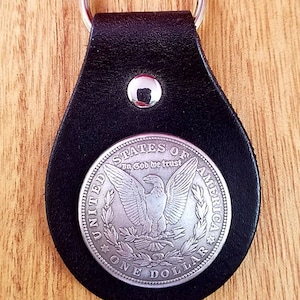May include: A silver dollar coin keychain with a black leather strap and a silver ring. The coin features an eagle with outstretched wings and the words "United States of America" and "In God We Trust".